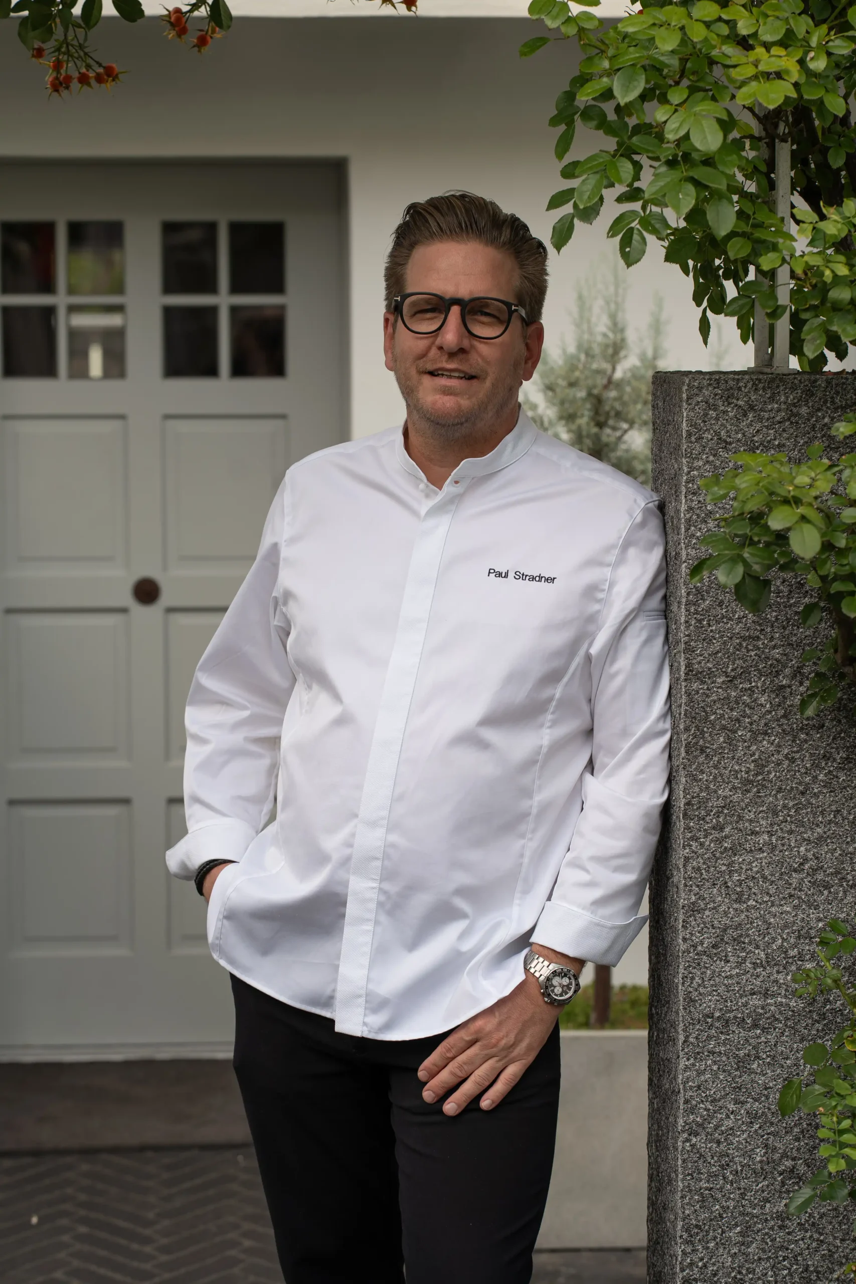 Porträt von Paul Stradner im weißen Kochjackett vor dem Malerhaus in Achern, stehend an einer Steinmauer mit Pflanzen.